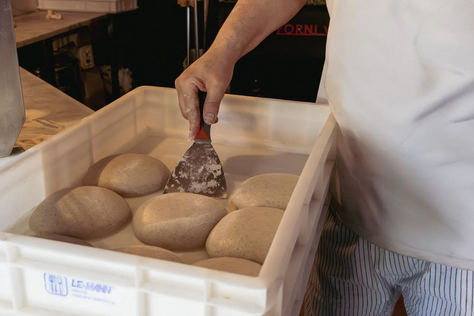 Una mano stacca con la spatola i panetti di impasto da una cassetta bianca.