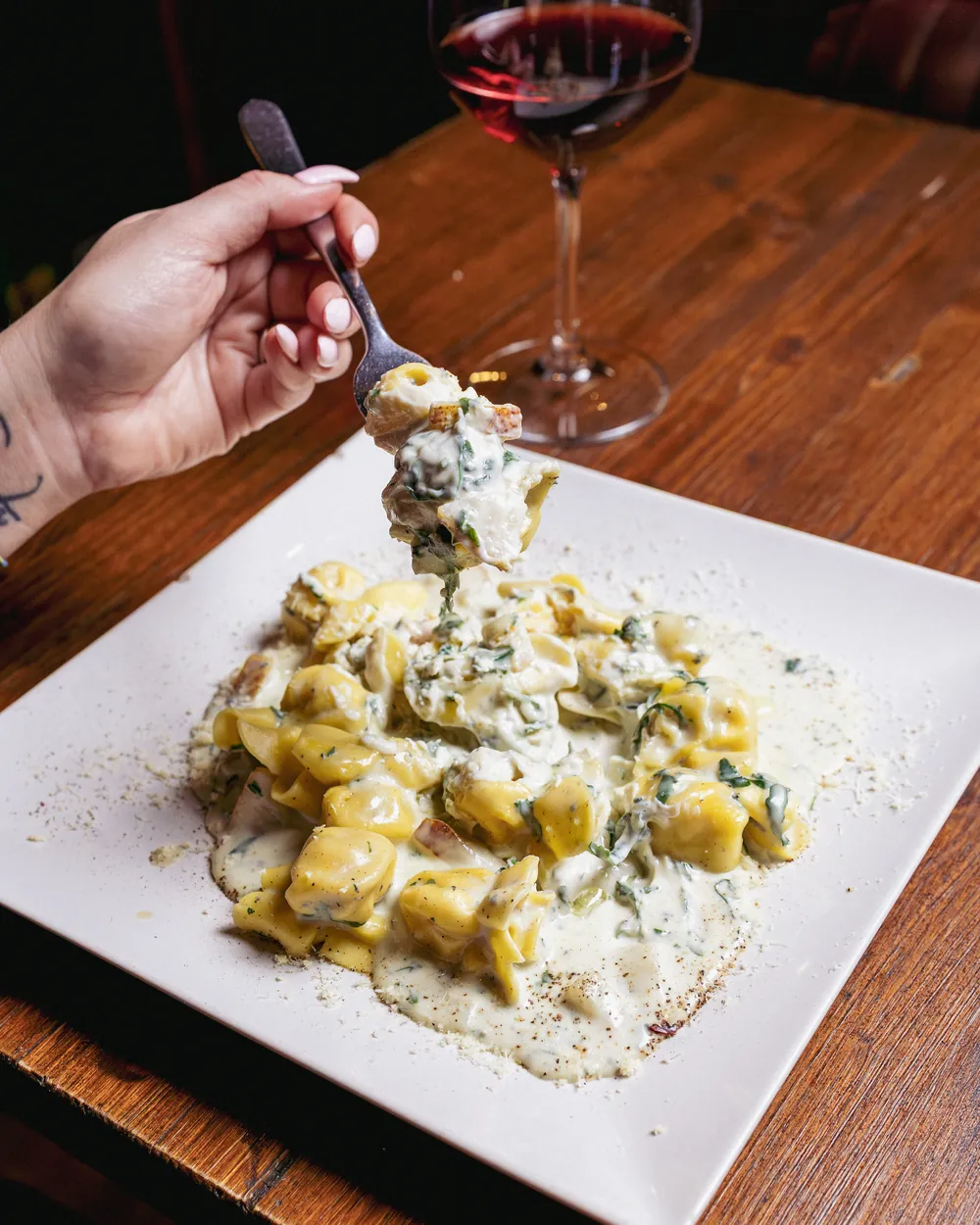 Fagottini in crema di gorgonzola con foglie verdi. Una forchetta ne solleva uno, dietro c’è un calice di vino rosso.