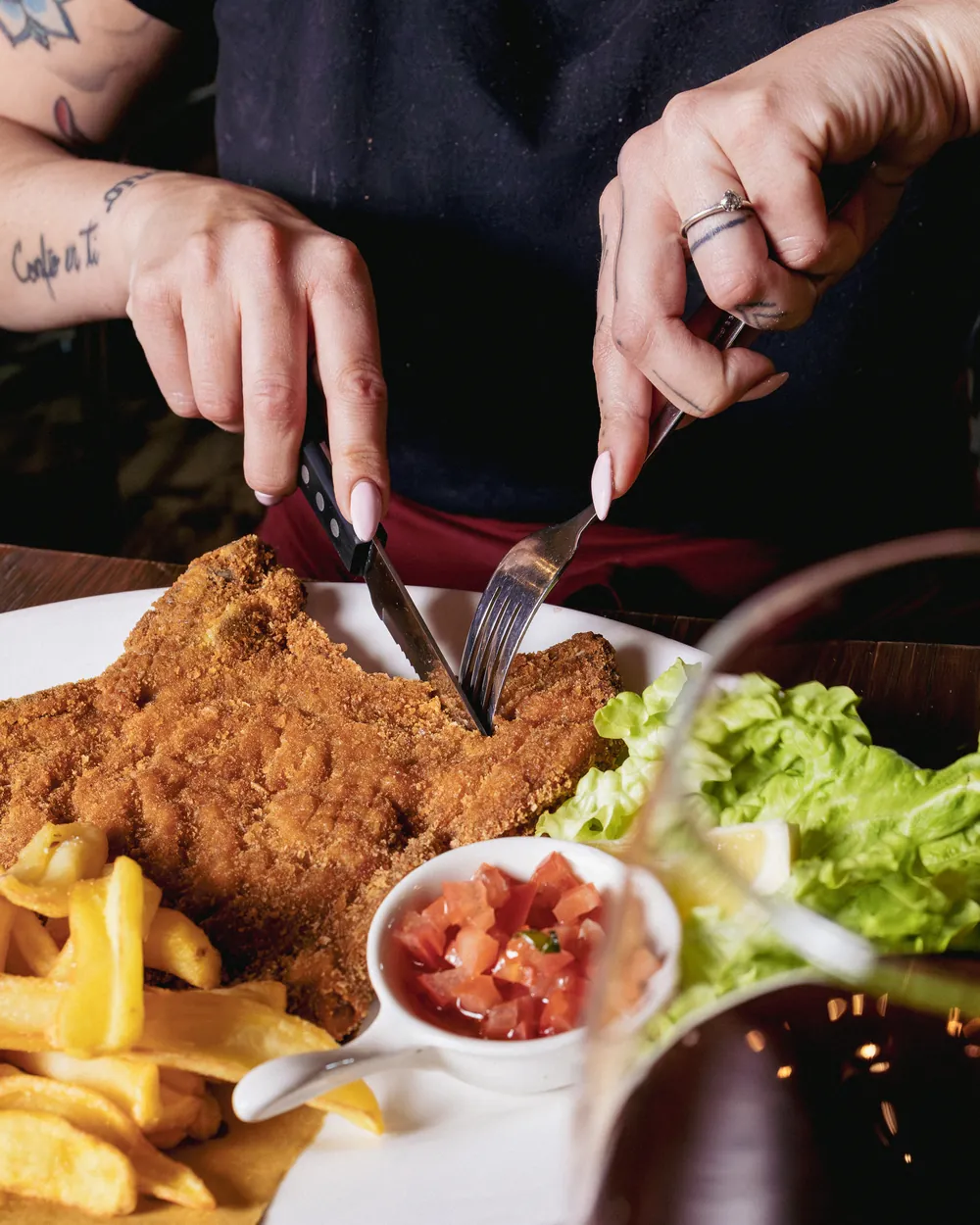 Due mani tagliano una milanese di vitella servita con patate fritte, insalata e una ciotolina di pomodoro a cubetti.