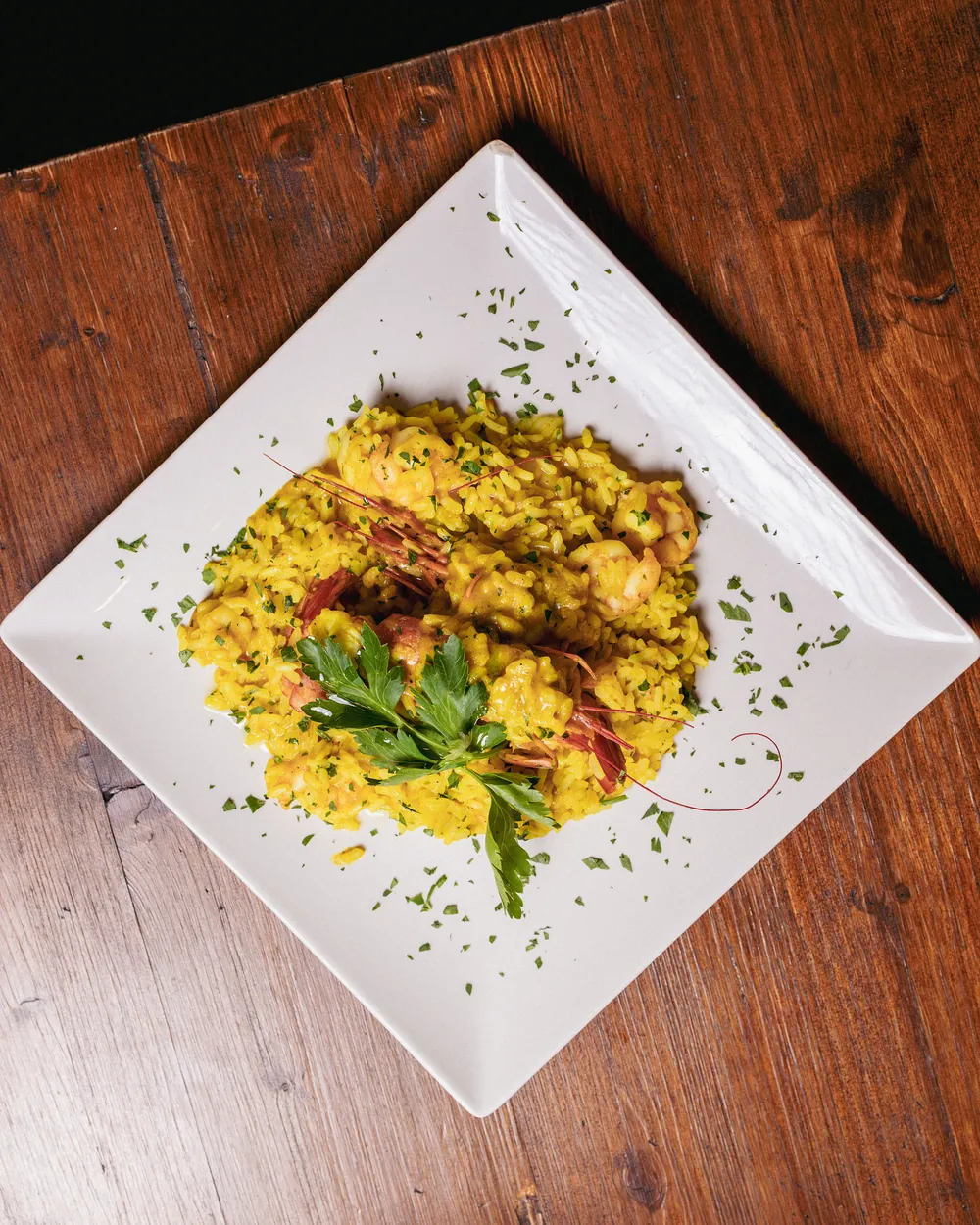 Risotto giallo al curry con gamberoni e prezzemolo, in un piatto quadrato bianco su un tavolo di legno scuro.